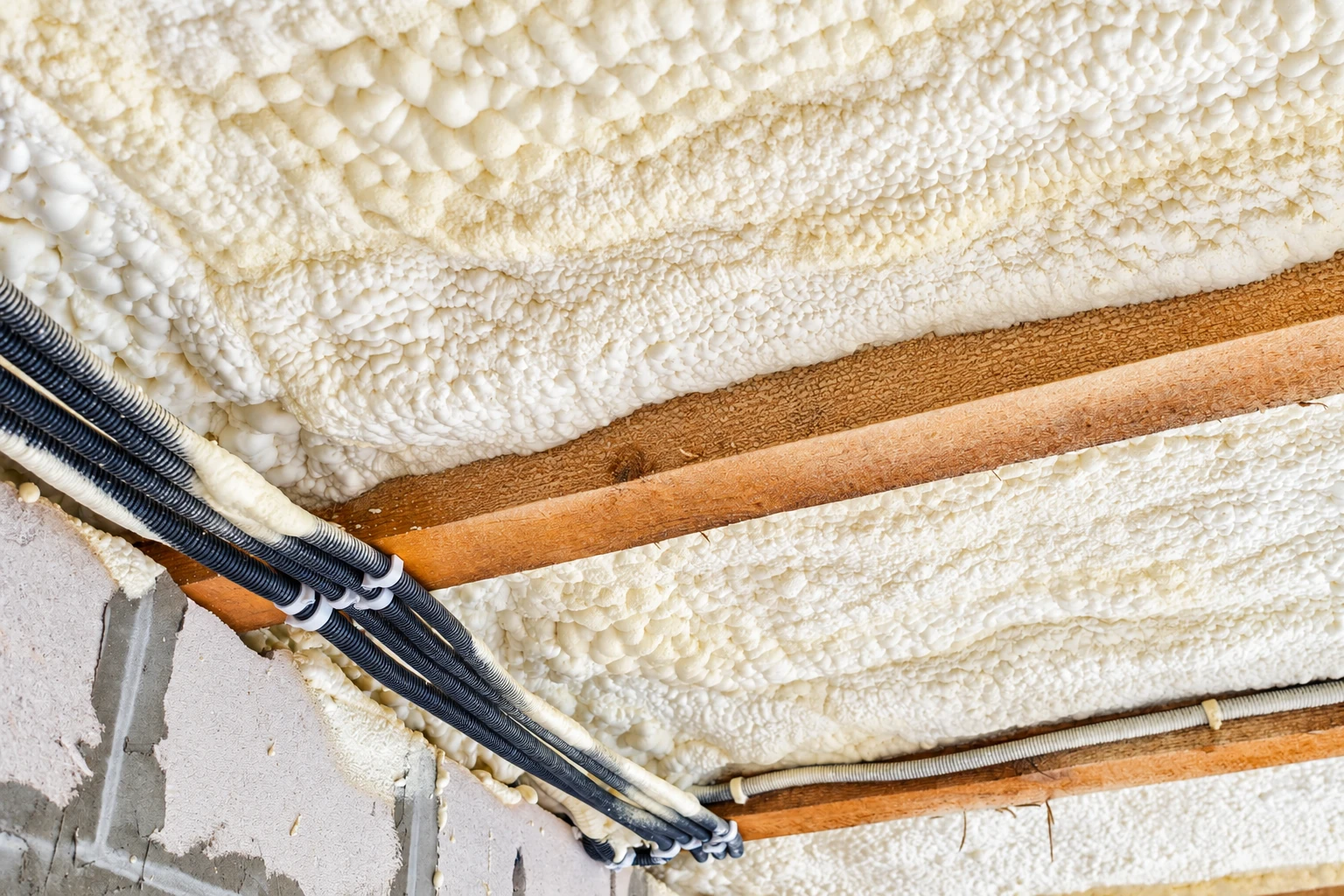 Close-up of thick white spray foam insulation between attic rafters, beside bundled black cables.