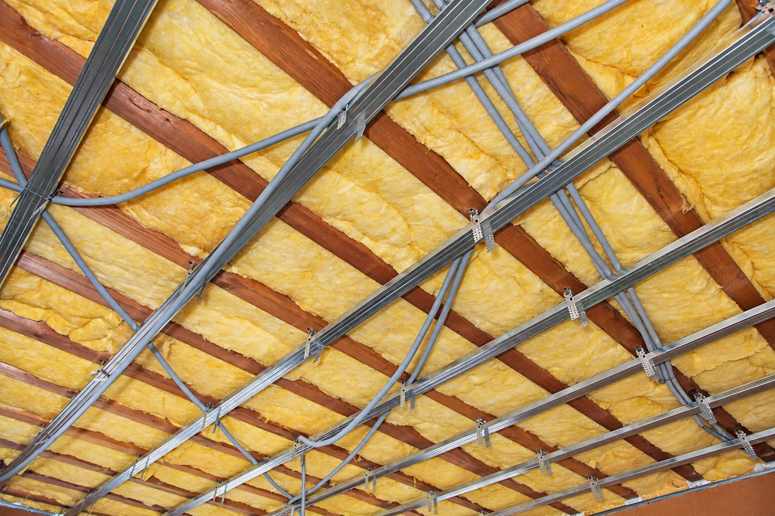Yellow fiberglass batts fill the spaces between roof rafters, crossed by metal framing channels and gray electrical conduits.