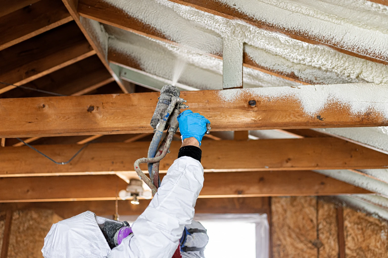A worker in protective gear sprays foam insulation onto the underside of a wooden roof structure.