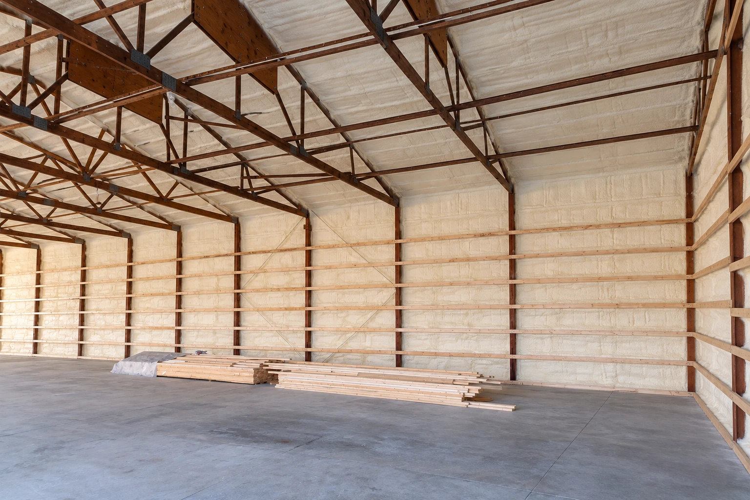 Commercial building interior with white spray foam insulation covering the roof and walls, exposed wooden trusses, and lumber stacked on the floor.